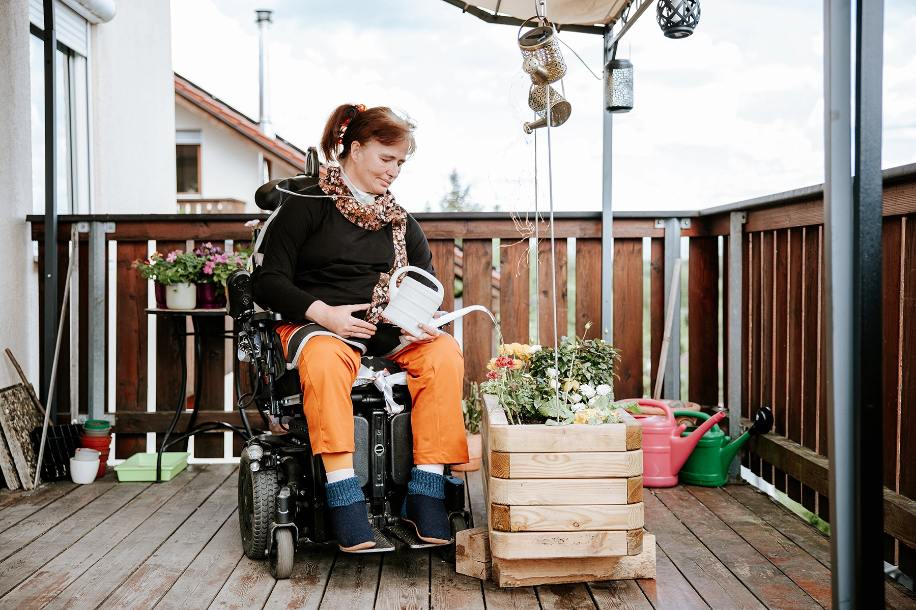 Person im Rollstuhl gießt Blumen auf einer Terrasse und sitzt neben einem Pflanzenkasten mit bunten Blüten.