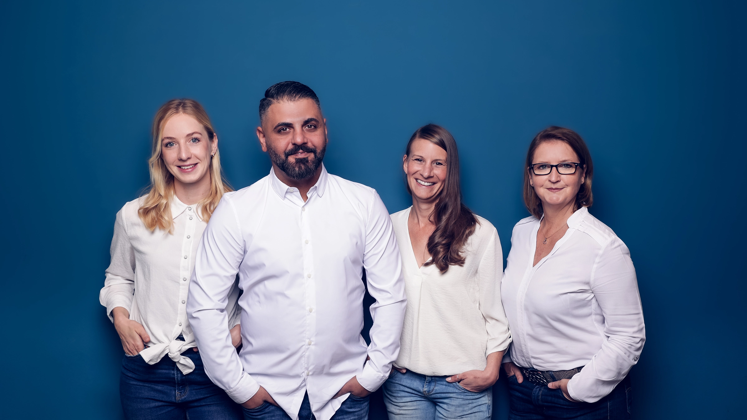 Drei freundlich lächelnde Frauen und ein Mann in weißen Hemden stehen zusammen vor blauem Hintergrund.