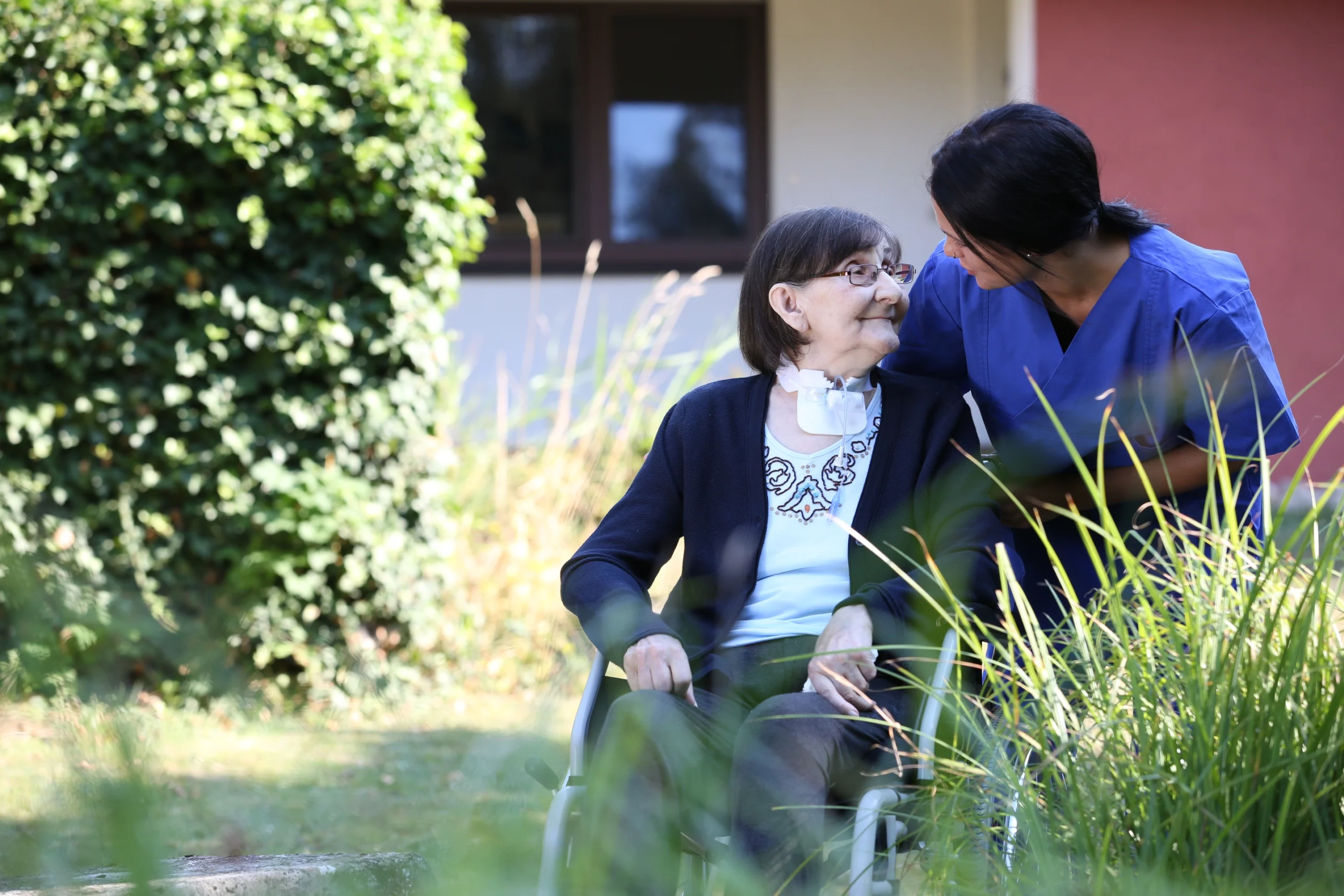 Pflegekraft betreut eine Person im Rollstuhl in einem Gartenbereich.