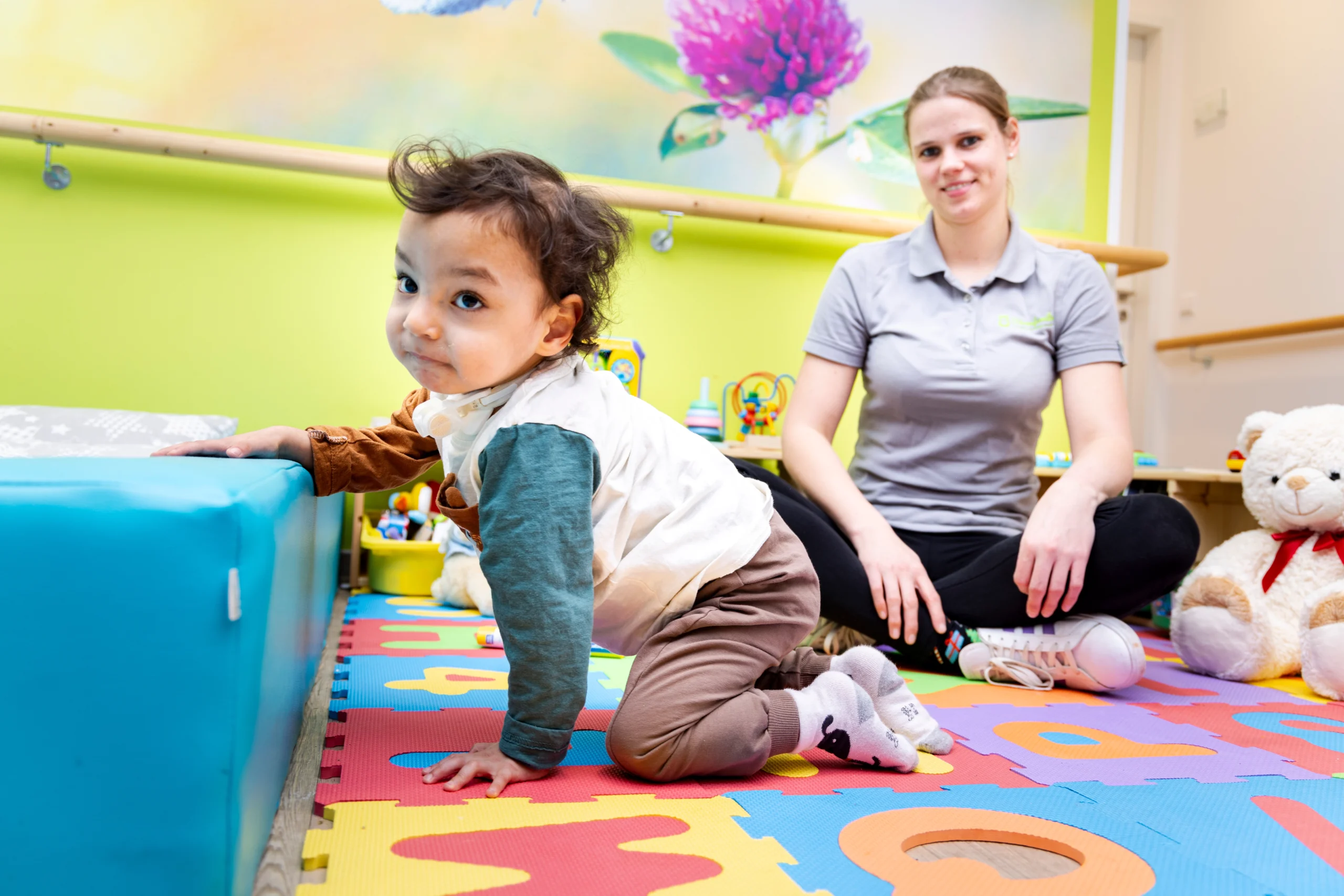 Kleinkind stützt sich auf einer bunten Spielmatte ab, während eine Therapeutin im Hintergrund sitzt und die Übung begleitet.