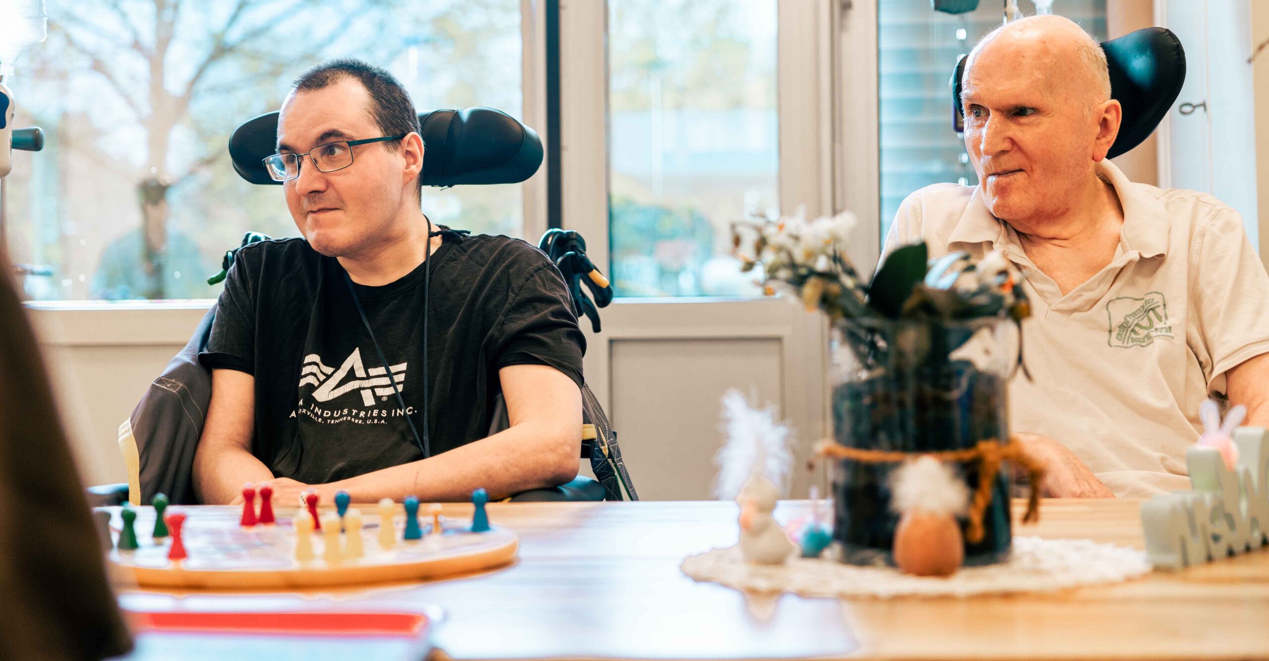 Zwei Personen sitzen in Rollstühlen an einem Tisch und spielen gemeinsam ein Brettspiel.