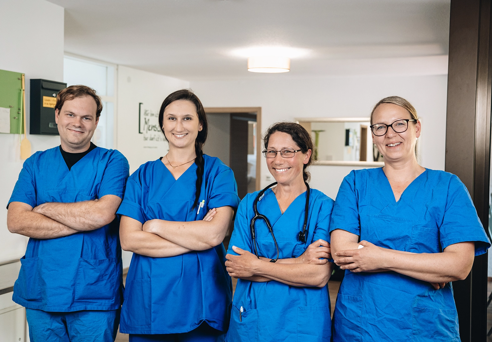 Vier Pflegekräfte in blauer Arbeitskleidung stehen nebeneinander mit verschränkten Armen in einem Innenbereich.