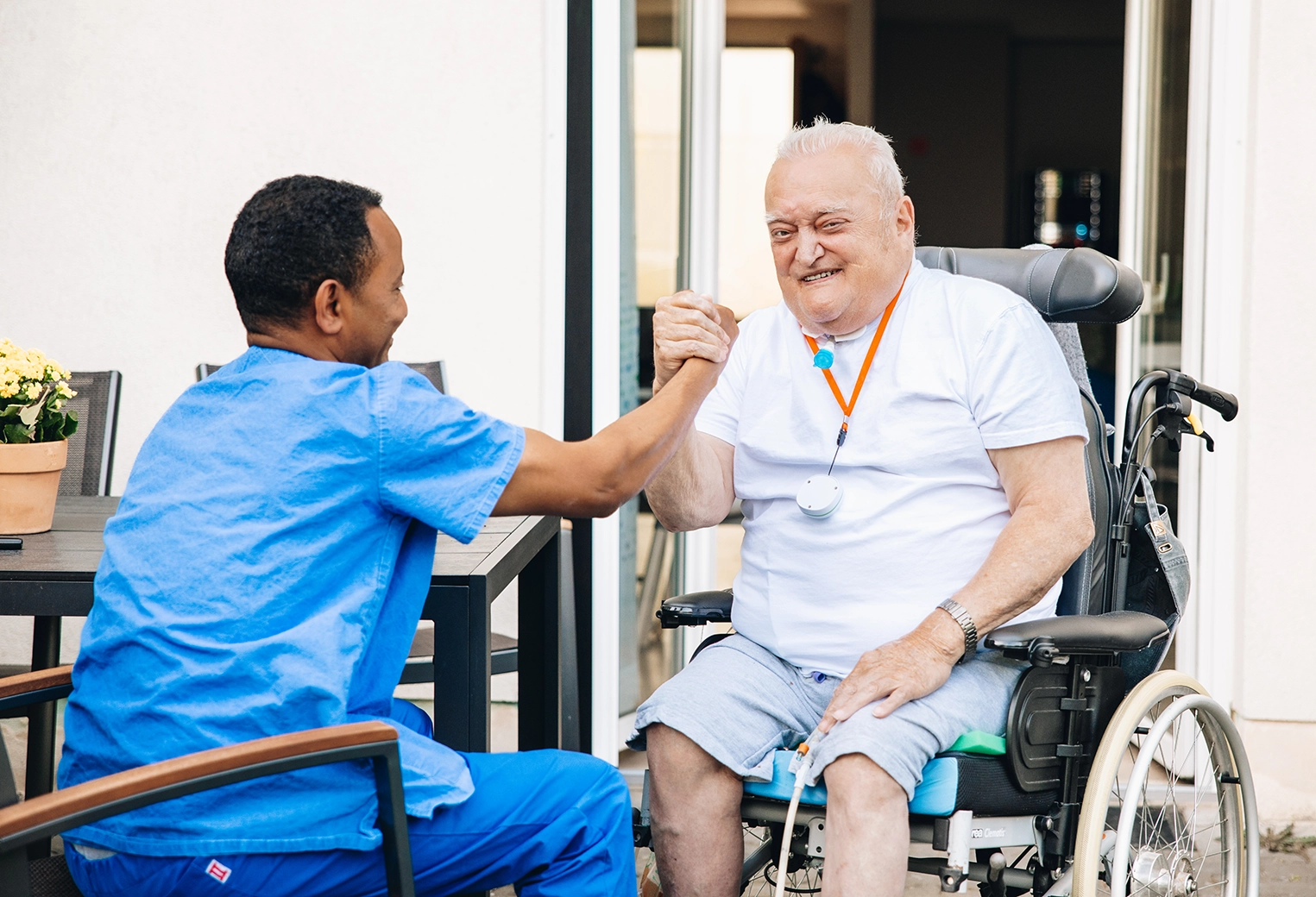 Pflegekraft begrüßt eine Person im Rollstuhl mit einem freundlichen Händedruck im Außenbereich.