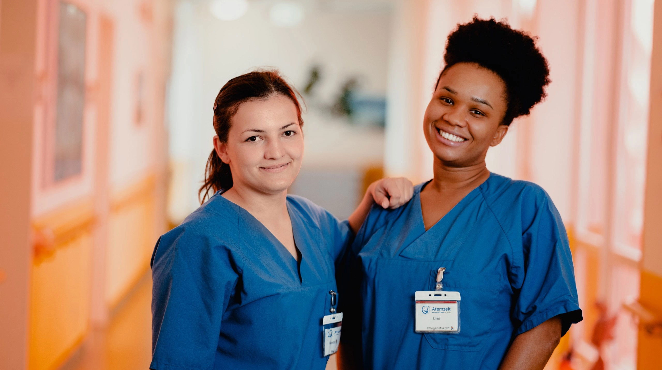 Zwei Pflegekräfte in blauen Arbeitskitteln stehen in einem hellen Krankenhausflur nebeneinander. Eine Person legt der anderen freundschaftlich den Arm auf die Schulter. Beide tragen Namensschilder.
