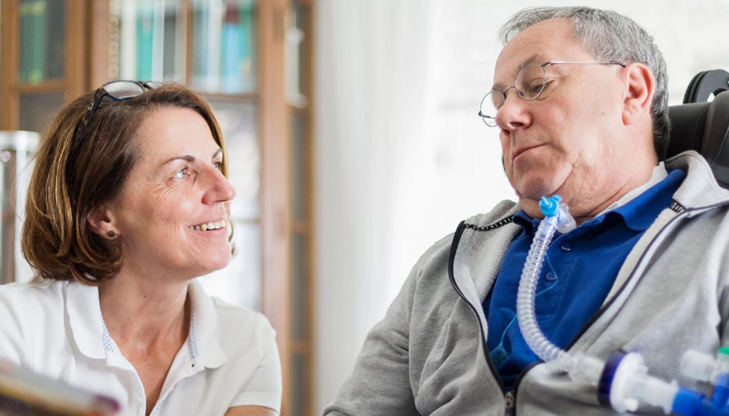 Pflegefachkraft sitzt im Gespräch mit beatmetem erwachsenem Patienten