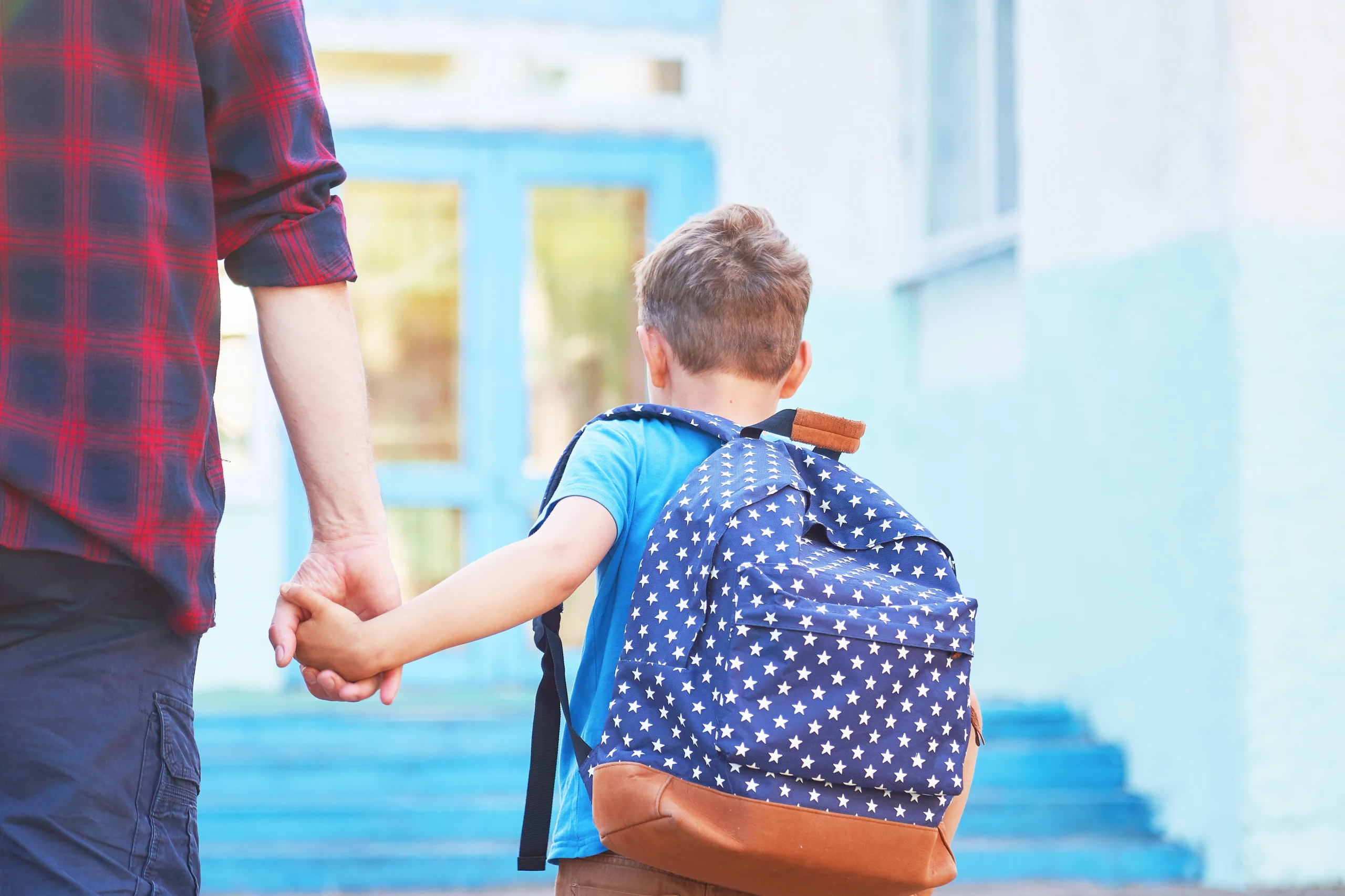 Kind mit Rucksack hält die Hand eines Erwachsenen auf dem Weg zur Schule.