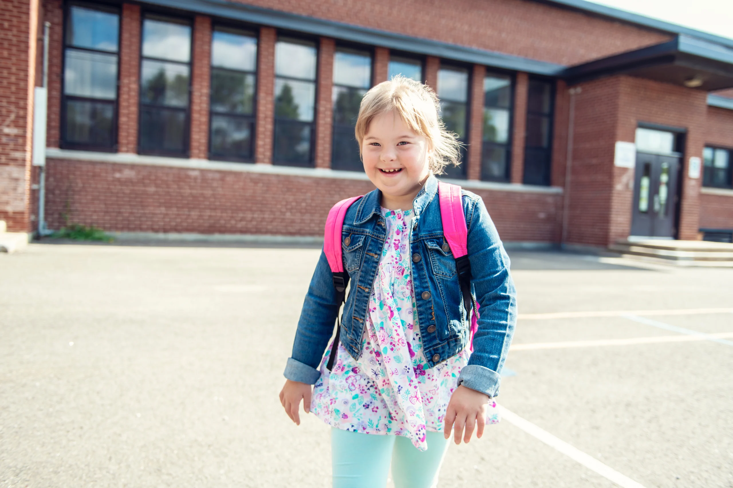 Mädchen mit Schulbegleitbedarf mit Rucksack steht lächelnd vor einem Schulgebäude auf dem Pausenhof.