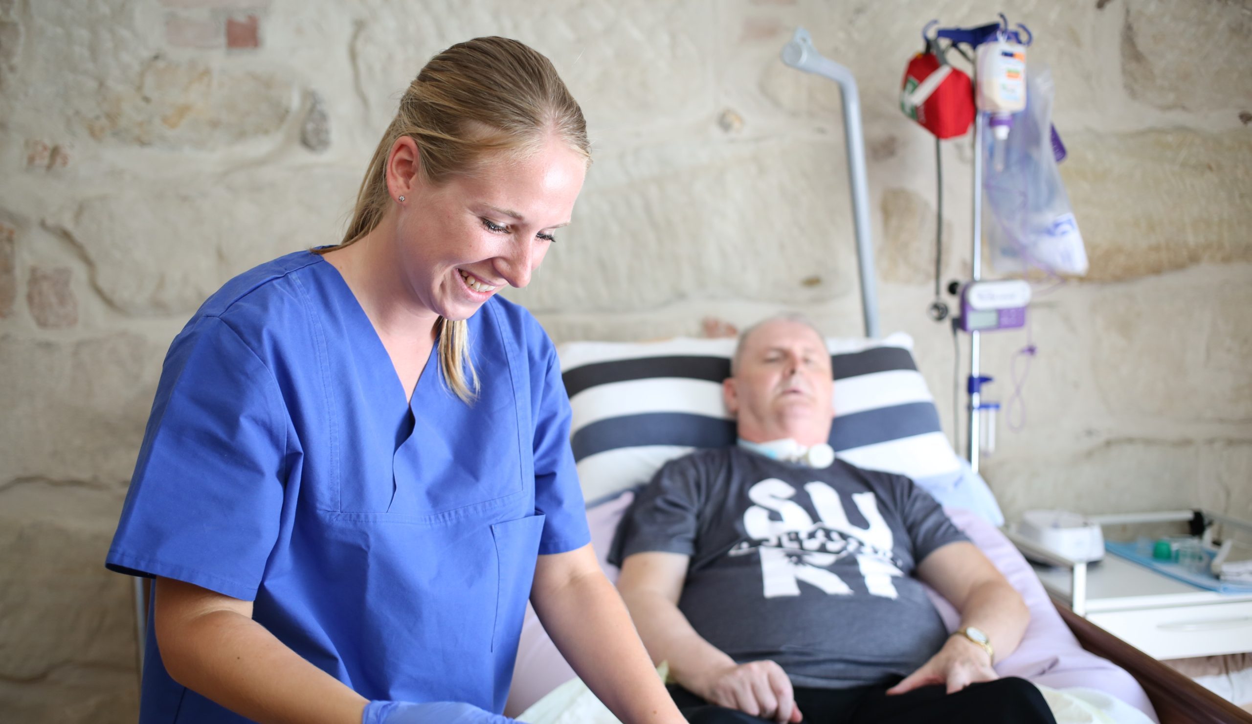 Pflegefachkraft in blauer Arbeitskleidung versorgt eine liegende Person in häuslicher Umgebung, im Hintergrund sind Ernährungsbeutel und medizinisches Equipment sichtbar.